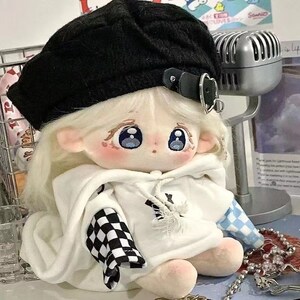 May include: A plush doll wearing a white hoodie with a black and white checked sleeve and a blue and white checked sleeve. The doll is wearing a black beret with a silver buckle. The doll has blonde hair and large blue eyes.
