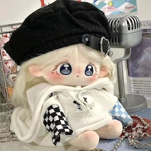 May include: A plush doll wearing a white hoodie with a black and white checked sleeve and a blue and white checked sleeve. The doll is wearing a black beret with a silver buckle. The doll has blonde hair and large blue eyes.
