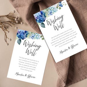 Könnte beinhalten: Zwei weiße Karten mit blauen Blumenmotiven und den Worten "Wishing Well" in eleganter Schrift. Die Karten enthalten Text über die Spende für eine Hochzeits-Wunschbox. Die Namen "Monika & Steven" sind unten aufgedruckt.