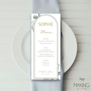 May include: A wedding menu card with the name "Sophie" in gold script. The menu lists starter, main, and dessert courses. Floral illustrations frame the text. The card is on a white plate and a blue napkin.