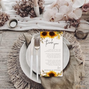 May include: A wedding reception table setting with a white plate, silverware, and a "thank you" card. The card features sunflowers and text that reads "Thank You" and a personalized message. The table is decorated with flowers.