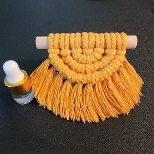 May include: A handmade macrame wall hanging in a warm yellow-orange hue, suspended from a natural wooden dowel. The design features a half-circle pattern with a fringe at the bottom. A small glass bottle with a white cap and gold trim is next to the macrame.