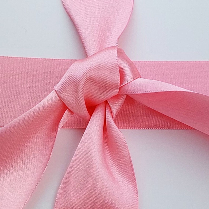 May include: A smooth, light pink satin ribbon tied into a simple knot. The ribbon features a lustrous, reflective surface with a consistent texture and a soft, pastel pink hue. The knot is positioned centrally, with two loose ends extending downwards.