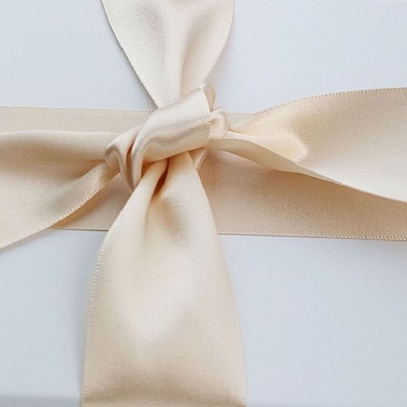 May include: A smooth, champagne-colored satin ribbon tied in a simple knot with two long, flowing tails extending downward and two shorter loops extending upward and to the sides, creating a decorative bow against a plain white surface.