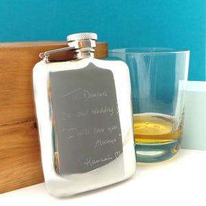 May include: A silver hip flask with a screw-on cap, engraved with a handwritten message. A clear glass with amber liquid sits beside the flask. The flask is rectangular with rounded edges, reflecting the surrounding light.