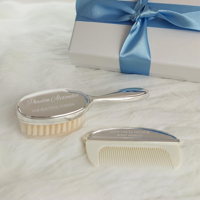 May include: A silver-toned baby hairbrush and comb set with white bristles and teeth. The brush is engraved with Phaedon Alexander and OUR BEAUTIFUL GODSON. The comb is engraved with LOVE UNCLE PHILIP & AUNT MARGO. A white gift box with a blue ribbon is included.