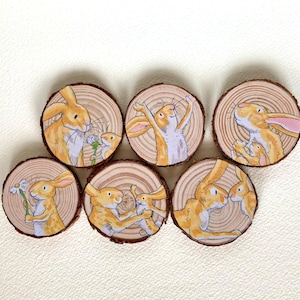 May include: A collection of seven circular wooden slices, each displaying a watercolour illustration of rabbits. The artwork depicts parent and child rabbits in various poses, using orange, white, and brown hues. The wood slices have a natural, rustic appearance.