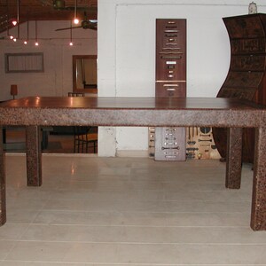 May include: A large, rectangular, brown wooden table with a textured surface. The table has four legs and is sitting on a light-colored floor.