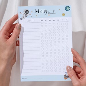 Puede incluir: Una lista de verificación imprimible azul y blanca con un diseño de temática espacial. La lista de verificación tiene un título que dice "Mein Raumschiffplaner" e incluye una sección para "Aufgabe" y los días de la semana. Hay círculos para marcar las tareas.