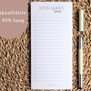 May include: A white shopping list notepad with horizontal lines and a row of circles for checkboxes. The notepad has the word "EINKAUFEN Shopping" at the top. A light green pen with a silver tip rests beside the notepad.