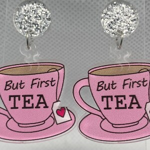 May include: Dangle earrings with a pink teacup design and the text "But First TEA". Each earring has a silver glitter ball and a tea bag with a heart. The teacups are on saucers.