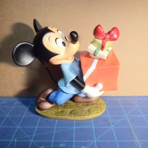 May include: A Mickey Mouse figurine carrying gifts. Mickey is dressed in a blue shirt and trousers, with white gloves. He holds a stack of presents, including a large orange box and smaller green boxes with red bows. The figurine is on a yellow base.