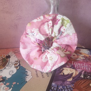 Peut inclure: Un chouchou en tissu rose et blanc avec un motif floral. Le chouchou est posé sur un livre dont la couverture porte le titre "Timeless Roma".