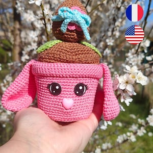 Könnte beinhalten: Ein gehäkelter rosa Hasentopf mit einem gehäkelten braunen Osterei oben drauf. Der Hasentopf hat eine rosa Herznase und schwarze Augen.