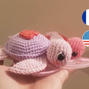 Peut inclure: Deux jouets en forme de tortue en crochet, en fil rose et violet. Les tortues ont des yeux noirs et l'une a un cœur rose sur sa carapace.