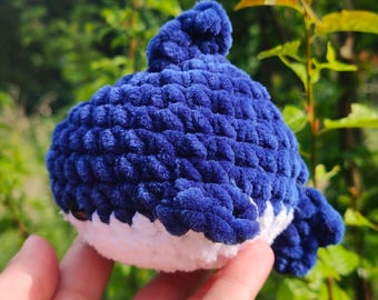 May include: A handmade, plush toy whale in shades of blue and white. The whale is crocheted with a textured yarn, featuring a rounded body and a small fin. The toy is held against a backdrop of green foliage.