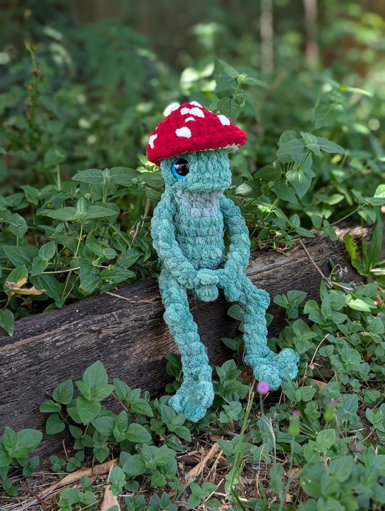 Pu&ograve; includere: Una creatura a forma di fungo fatta a mano all'uncinetto, con un cappello rosso a pois bianchi, corpo blu e grandi occhi blu. Il peluche &egrave; seduto su un tronco circondato da verde. Ha gambe e braccia lunghe.