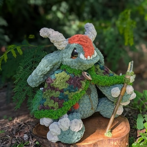 Peut inclure: Une créature en peluche avec une fourrure vert sauge, de grandes oreilles et deux cornes, arborant une tache de fourrure orange sur la tête. Elle porte une cape à motif camouflage fixée par un bouton en bois et tient un bâton en bois surmonté d'un cristal.