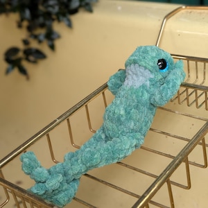 Pu&ograve; includere: Un peluche a forma di delfino all'uncinetto turchese con grandi occhi blu e pancia grigia. Il peluche &egrave; appoggiato su una griglia metallica dorata. Lo sfondo &egrave; giallo tenue.