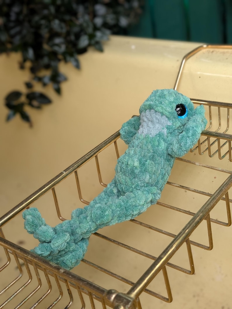 Pu&ograve; includere: Un peluche a forma di delfino all'uncinetto turchese con grandi occhi blu e pancia grigia. Il peluche &egrave; appoggiato su una griglia metallica dorata. Lo sfondo &egrave; giallo tenue.