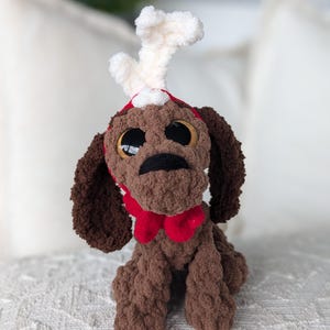 May include: A brown crocheted reindeer plush toy with big eyes, a red scarf, and white antlers. The toy is sitting on a white surface.