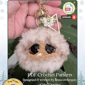 May include: A plush keychain head with the text "Arki Jr. Keychain Head". The keychain has a fluffy, light brown exterior, large black eyes, and a small mouth. A gold-colored charm with a butterfly design is attached.