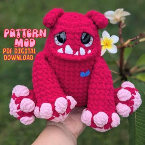 May include: A crocheted plush toy monster with pink feet, a blue bow, and large black eyes. The monster is sitting on a hand with a green background.