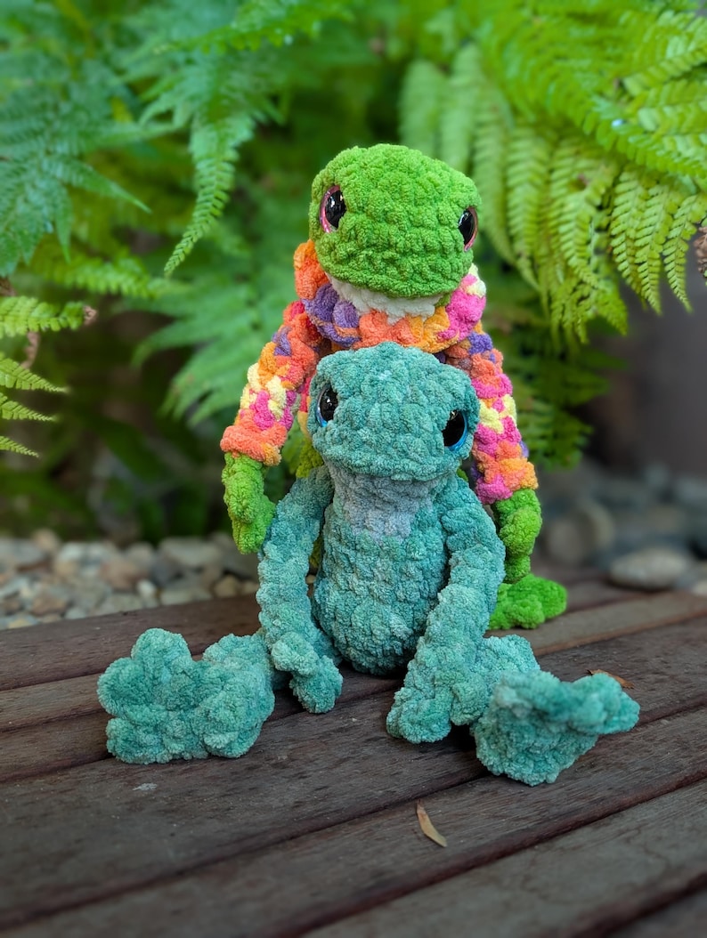 Pu&ograve; includere: Due peluche a forma di rana fatti a mano all'uncinetto. Uno &egrave; verde con un maglione colorato, l'altro &egrave; verde acqua. Le rane hanno grandi occhi neri e sono sedute su una superficie di legno. Felci sullo sfondo.