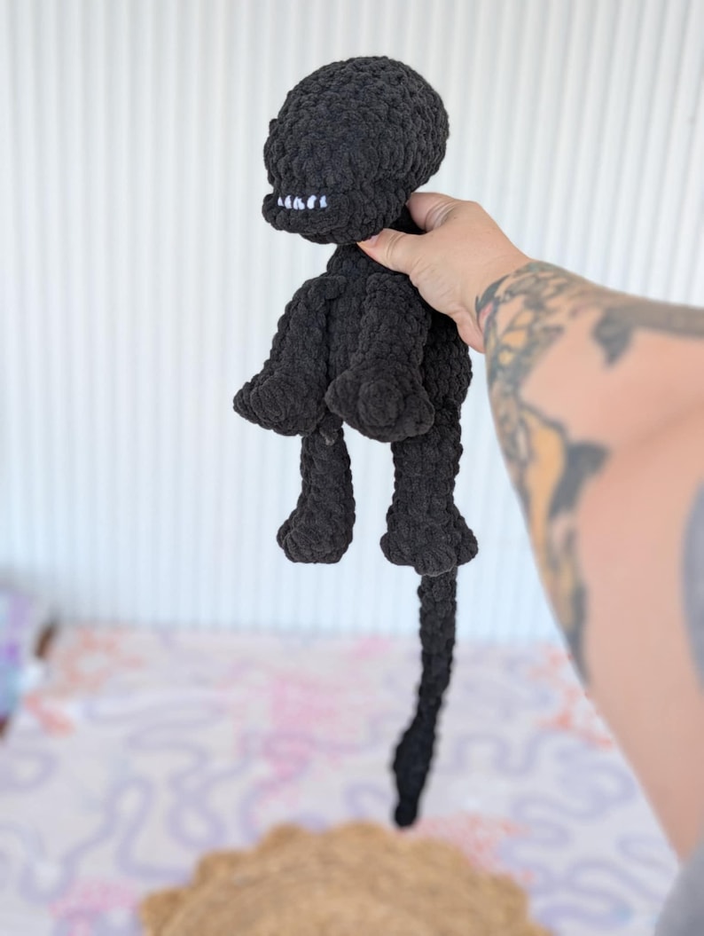 May include: A handmade black crochet doll with a round head, small arms, and legs. The doll has a stitched mouth with white teeth and a long tail. The doll is held up against a white background.