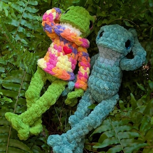 Pu&ograve; includere: Due peluche a forma di rana fatti a mano all'uncinetto riposano tra felci verdi. Una rana &egrave; verde con un maglione colorato e un cuore rosso. L'altra rana &egrave; blu. Entrambe hanno grandi occhi scuri e sono realizzate in filato morbido e strutturato.