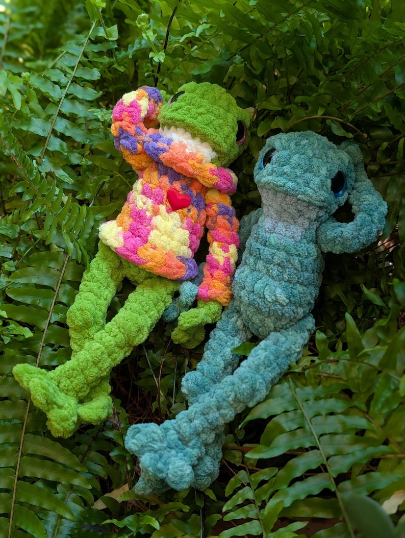 Pu&ograve; includere: Due peluche a forma di rana fatti a mano all'uncinetto riposano tra felci verdi. Una rana &egrave; verde con un maglione colorato e un cuore rosso. L'altra rana &egrave; blu. Entrambe hanno grandi occhi scuri e sono realizzate in filato morbido e strutturato.