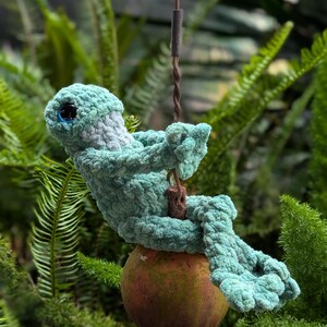 Pu&ograve; includere: Una creatura all'uncinetto turchese fatta a mano con occhi blu, appollaiata su un frutto marrone. La figura di peluche &egrave; appesa a un filo, su uno sfondo di fogliame verde. Il design &egrave; stravagante e fatto a mano. Il frutto misura circa 7 cm di diametro.