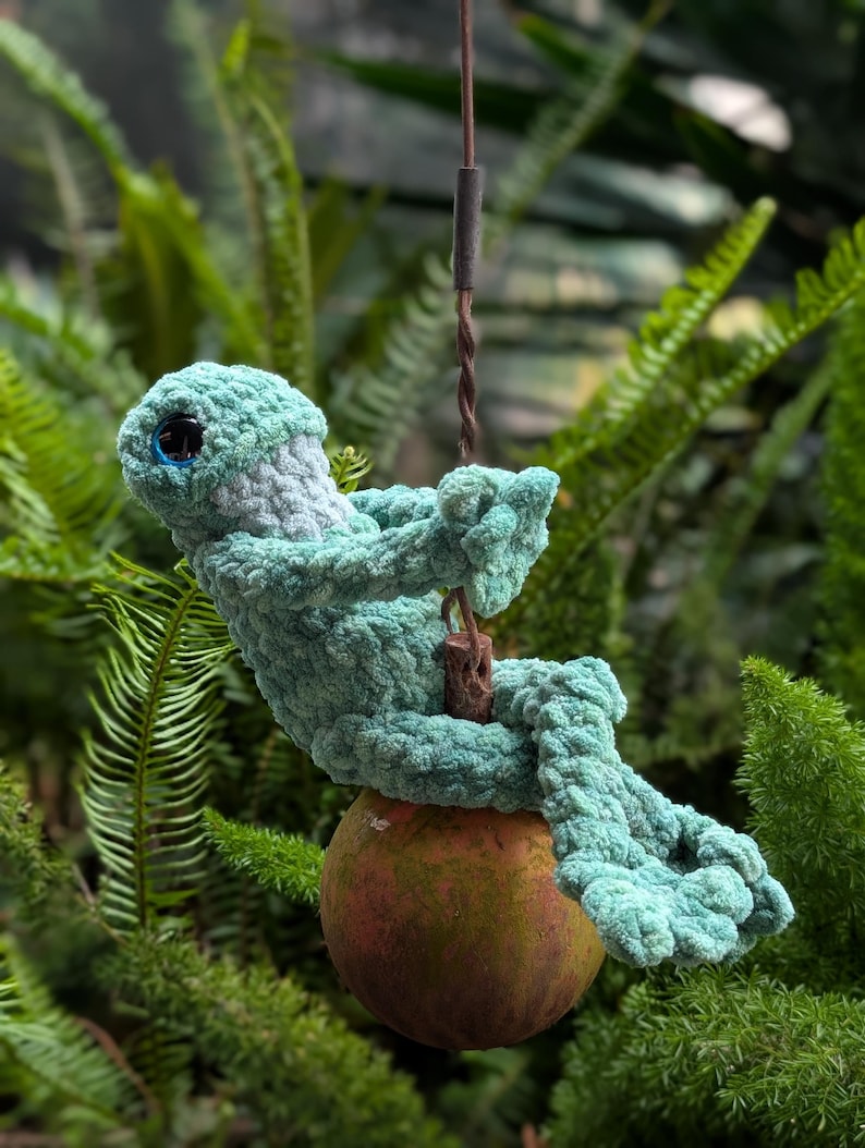 Pu&ograve; includere: Una creatura all'uncinetto turchese fatta a mano con occhi blu, appollaiata su un frutto marrone. La figura di peluche &egrave; appesa a un filo, su uno sfondo di fogliame verde. Il design &egrave; stravagante e fatto a mano. Il frutto misura circa 7 cm di diametro.