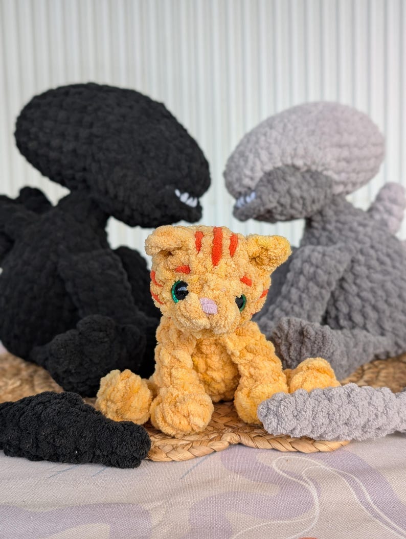 May include: A handmade crochet plushie scene featuring a small orange tabby cat with red stripes, flanked by two large, dark gray and light gray alien figures. The toys are on a woven mat.