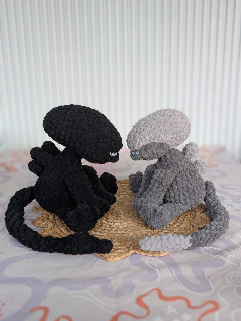 May include: Two crocheted alien plush toys. One is black, the other is gray. Both have elongated heads, small arms, and long tails. They are sitting on a woven mat, with a patterned background.