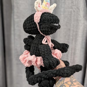 May include: A handmade black crochet creature wearing a pink tutu, a pink headband, and a unicorn horn with pastel accents. The plush toy has a small mouth and is held up by a person.