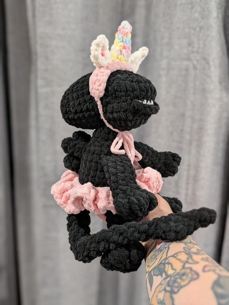 May include: A handmade black crochet creature wearing a pink tutu, a pink headband, and a unicorn horn with pastel accents. The plush toy has a small mouth and is held up by a person.