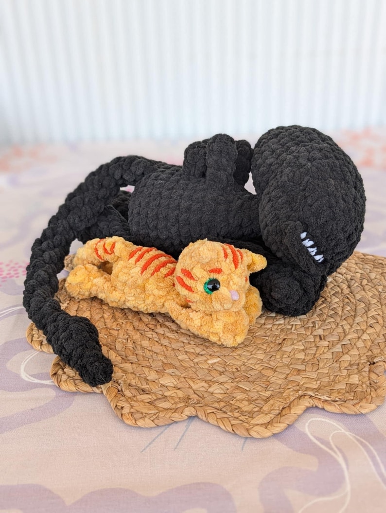 May include: A handmade crochet cat and a black creature rest on a woven placemat. The orange cat has red stripes and green eyes. The black creature has a long tail and a round head.