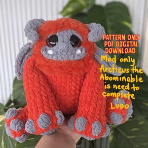 May include: A crocheted plush toy in the shape of a monster with orange fur, gray face, and large eyes. The toy is being held by a hand. The text "PATTERN ONLY PDF DIGITAL DOWNLOAD Mod only Arcticus the Abominable is need to Complete LUDO" is written in yellow on a white background.