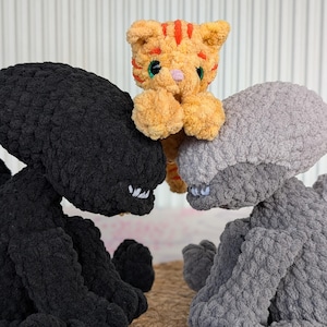 May include: A plush toy arrangement featuring a small orange tabby cat nestled between two large, textured alien figures. One alien is black, the other is gray. The cat has orange stripes and green eyes.