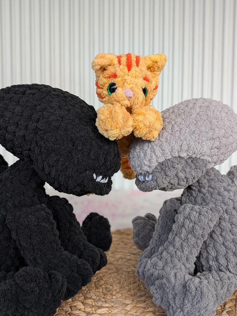 May include: A plush toy arrangement featuring a small orange tabby cat nestled between two large, textured alien figures. One alien is black, the other is gray. The cat has orange stripes and green eyes.