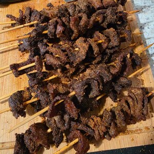 May include: Several wooden skewers threaded with thin, dark brown strips of seasoned meat. The meat appears cooked and coated in a coarse, granular spice rub, displaying a deep mahogany color with visible texture from the seasoning.