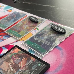 May include: A close-up of trading cards with the text "Jewelry Bonney" and "Cavendish". The cards are on a pink surface with a cartoon design. Two black oval tokens with the word "FREEZED" are also visible.