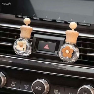 May include: Two clear glass car air fresheners with wooden tops and flower inserts. They are positioned on a car dashboard near air vents and control knobs. The car's dashboard is black.