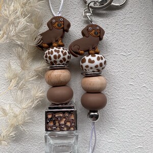 May include: A brown and white dachshund car charm with a brown and white patterned bead and a brown and wood bead. The charm is attached to a clear glass bottle with a silver top and a silver metal ring. The charm is also attached to a silver key ring.