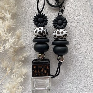 May include: Black and white cow print car air freshener with black beads and black flower charms. The air freshener is attached to a black string with a silver metal ring.