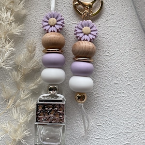 May include: Two beaded car air fresheners. Each has a gold keyring, a lavender daisy, wooden beads, gold rings, lavender and white beads, and a clear glass bottle with a decorative top. One has a white string, the other a gold tassel.