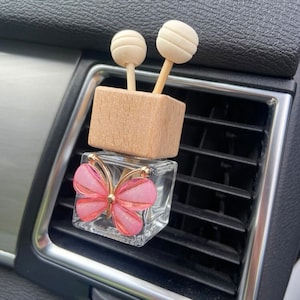 May include: A pink butterfly car air freshener with a wooden top and two white diffuser sticks. The air freshener is attached to a car's air vent.