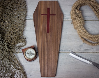 Könnte beinhalten: Ein Holzsarg mit einem roten Kreuz auf der Oberseite, umgeben von einem Seil, einem Kompass und einem Fischernetz auf einem weißen Holzgrund.