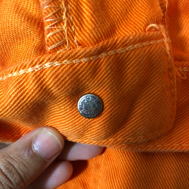 Op de afbeelding: Close-up van een feloranje denim stof met een zilveren metalen knoop. De knoop heeft het woord "GOLD" erop gegraveerd.